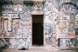 A wall covered with an intricate and vibrant mural featuring abstract and geometric shapes, faces, and artistic doodles. The artwork is predominantly in black and white with some colored elements and includes various patterns and symbols. In the center of the wall, there is an open doorway, creating a contrast between the mural and the dark interior beyond.