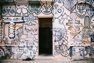 A wall covered with an intricate and vibrant mural featuring abstract and geometric shapes, faces, and artistic doodles. The artwork is predominantly in black and white with some colored elements and includes various patterns and symbols. In the center of the wall, there is an open doorway, creating a contrast between the mural and the dark interior beyond.