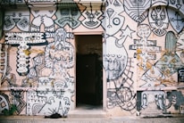 A wall covered with an intricate and vibrant mural featuring abstract and geometric shapes, faces, and artistic doodles. The artwork is predominantly in black and white with some colored elements and includes various patterns and symbols. In the center of the wall, there is an open doorway, creating a contrast between the mural and the dark interior beyond.
