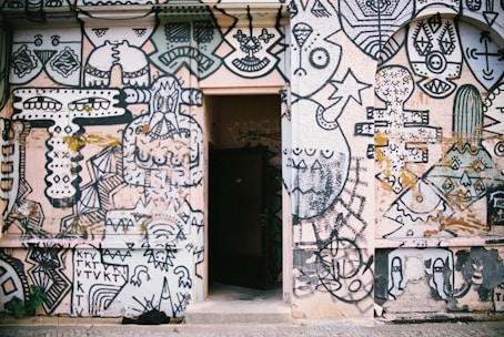 A wall covered with an intricate and vibrant mural featuring abstract and geometric shapes, faces, and artistic doodles. The artwork is predominantly in black and white with some colored elements and includes various patterns and symbols. In the center of the wall, there is an open doorway, creating a contrast between the mural and the dark interior beyond.