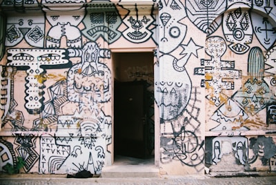 A wall covered with an intricate and vibrant mural featuring abstract and geometric shapes, faces, and artistic doodles. The artwork is predominantly in black and white with some colored elements and includes various patterns and symbols. In the center of the wall, there is an open doorway, creating a contrast between the mural and the dark interior beyond.