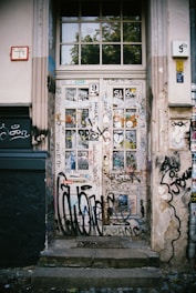 A wooden door covered in various colorful graffiti and stickers. The surrounding wall also features graffiti, giving the area an urban, gritty atmosphere. The door is set within a white and beige building facade, with a mix of modern and traditional architectural elements.