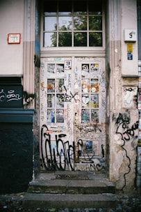 A wooden door covered in various colorful graffiti and stickers. The surrounding wall also features graffiti, giving the area an urban, gritty atmosphere. The door is set within a white and beige building facade, with a mix of modern and traditional architectural elements.