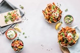 An arrangement of food on a light-colored surface features dishes with sweet potato fries topped with herbs, avocado, and other vegetables. A small bowl of dip, sliced lemons, and a cutting board with chopped vegetables are also present. Garnishes and herbs are scattered around for decoration.