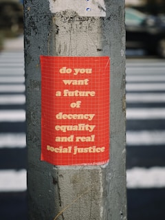 A red poster with white text is pasted on a weathered concrete pole. The background features a pedestrian crossing, suggesting an urban setting. The text on the poster promotes values of decency, equality, and social justice.