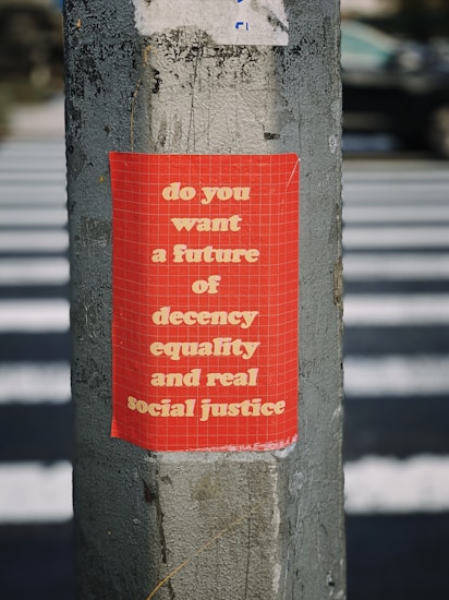 A red poster with white text is pasted on a weathered concrete pole. The background features a pedestrian crossing, suggesting an urban setting. The text on the poster promotes values of decency, equality, and social justice.