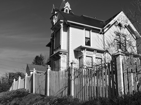 A large Victorian-style house with intricate woodwork on the eaves is situated behind a tall, wooden picket fence. The scene is captured in black and white, highlighting the architectural details and the bare branches of trees surrounding the property.
