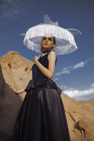 A woman wearing a dark, elegant dress is set against a desert landscape with sandy rock formations. Her large, unique hat with a wide brim adorned with fringe adds a dramatic effect. The sky is clear and blue, enhancing the serene and stylish scene.