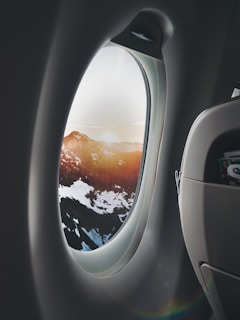 A view through an airplane window shows a breathtaking landscape of snow-capped mountains bathed in warm sunlight. The interior of the airplane, including part of a seat and window frame, is visible, adding a sense of travel and adventure.