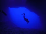 The silhouetted figure of a scuba diver exploring a sunlit ocean cave.