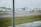 A runway scene at an airport with two small aircraft parked on the tarmac. The area is lined with orange traffic cones and yellow signage. In the background, there are large hangars and some distant buildings. The day appears to be overcast, and a patch of grass is visible beside the runway.