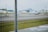 A runway scene at an airport with two small aircraft parked on the tarmac. The area is lined with orange traffic cones and yellow signage. In the background, there are large hangars and some distant buildings. The day appears to be overcast, and a patch of grass is visible beside the runway.