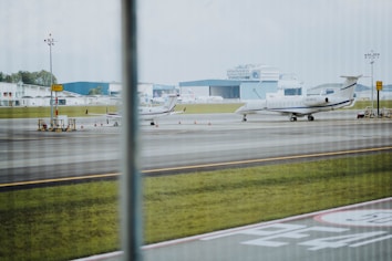 A runway scene at an airport with two small aircraft parked on the tarmac. The area is lined with orange traffic cones and yellow signage. In the background, there are large hangars and some distant buildings. The day appears to be overcast, and a patch of grass is visible beside the runway.