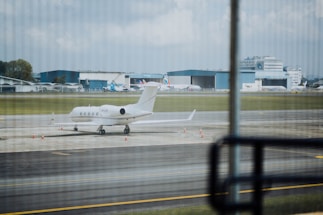 A private jet is parked on an airport runway with several cones placed around it. In the background, there are large hangars and more commercial airplanes. The scene is observed from behind glass windows, creating a slightly striped pattern across the view.