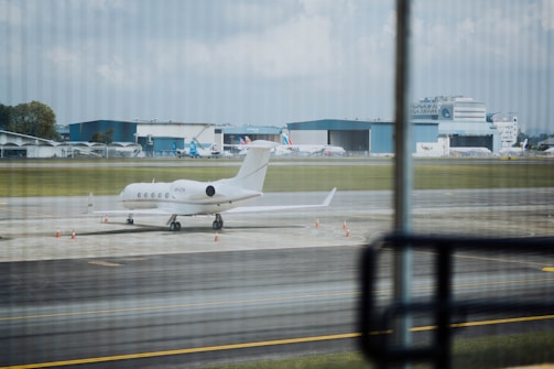 A private jet is parked on an airport runway with several cones placed around it. In the background, there are large hangars and more commercial airplanes. The scene is observed from behind glass windows, creating a slightly striped pattern across the view.