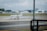 A private jet is parked on an airport runway with several cones placed around it. In the background, there are large hangars and more commercial airplanes. The scene is observed from behind glass windows, creating a slightly striped pattern across the view.