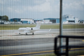 A private jet is parked on an airport runway with several cones placed around it. In the background, there are large hangars and more commercial airplanes. The scene is observed from behind glass windows, creating a slightly striped pattern across the view.