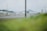 A view of an airport tarmac with two small private jets parked. In the background, there are industrial hangars and structures. The foreground is slightly blurred with green grass, possibly indicating the photo was taken through a fence or from a distance.