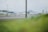 A view of an airport tarmac with two small private jets parked. In the background, there are industrial hangars and structures. The foreground is slightly blurred with green grass, possibly indicating the photo was taken through a fence or from a distance.