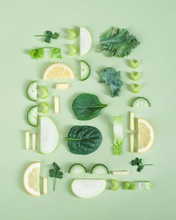 An arrangement of sliced fruits and vegetables, including lemon, cucumber, celery, apple, kale, and spinach, is placed on a light green background. The pieces are symmetrically organized, creating an aesthetically pleasing pattern with different shades of green and yellow.