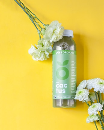 A green bottle labeled 'DOSE ORGANIC' with the word 'cactus' on it is placed on a bright yellow background. Beside the bottle, there are several light green and white flowers, adding a fresh and natural element to the composition.