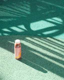 A bottle of smoothie is placed on a textured green surface with dramatic shadows cast by what appears to be foliage, creating dynamic patterns across the ground.