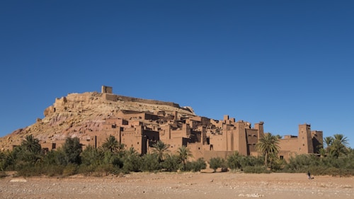 An ancient fortified village with earthen buildings is nestled against a rocky hillside. The structures are made of reddish-brown clay and have a rustic appearance. Palm trees and other green vegetation surround the settlement, contrasting with the clear blue sky above.