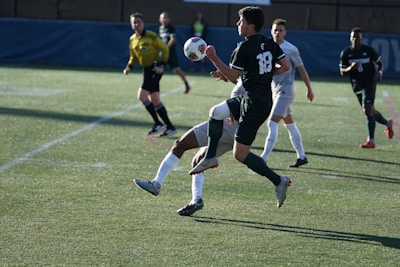 A soccer match taking place on a grassy field. Players are in action, with one player in black jersey number 18 controlling the ball mid-air while being challenged by an opposing player in a white jersey. A referee in a yellow uniform observes the game closely in the background, along with other players.