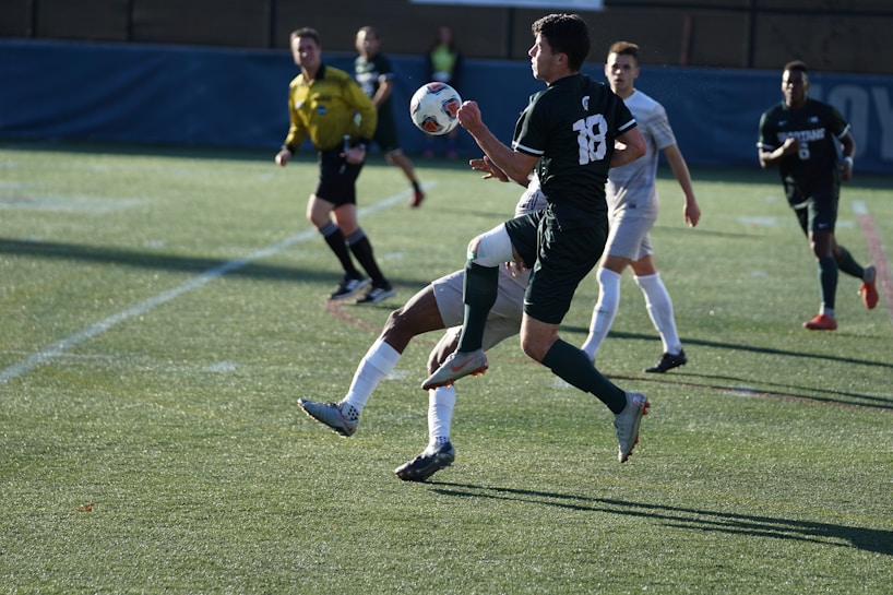 A soccer match taking place on a grassy field. Players are in action, with one player in black jersey number 18 controlling the ball mid-air while being challenged by an opposing player in a white jersey. A referee in a yellow uniform observes the game closely in the background, along with other players.