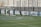 A soccer match on a field where players are in action near a goalpost. Several players in black uniforms are positioned in and around the goal area, while one player in white is executing an acrobatic move mid-air with a soccer ball nearby. The backdrop includes a tall wall with faded writing on it, and the field features green artificial turf with blue markings.