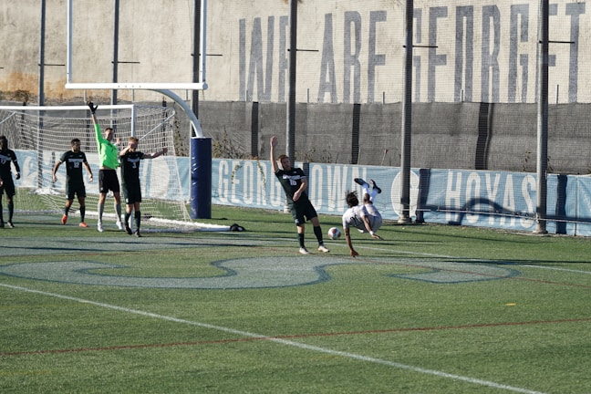 A soccer match on a field where players are in action near a goalpost. Several players in black uniforms are positioned in and around the goal area, while one player in white is executing an acrobatic move mid-air with a soccer ball nearby. The backdrop includes a tall wall with faded writing on it, and the field features green artificial turf with blue markings.