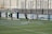 A soccer match on a field where players are in action near a goalpost. Several players in black uniforms are positioned in and around the goal area, while one player in white is executing an acrobatic move mid-air with a soccer ball nearby. The backdrop includes a tall wall with faded writing on it, and the field features green artificial turf with blue markings.