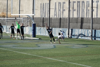 A soccer match on a field where players are in action near a goalpost. Several players in black uniforms are positioned in and around the goal area, while one player in white is executing an acrobatic move mid-air with a soccer ball nearby. The backdrop includes a tall wall with faded writing on it, and the field features green artificial turf with blue markings.