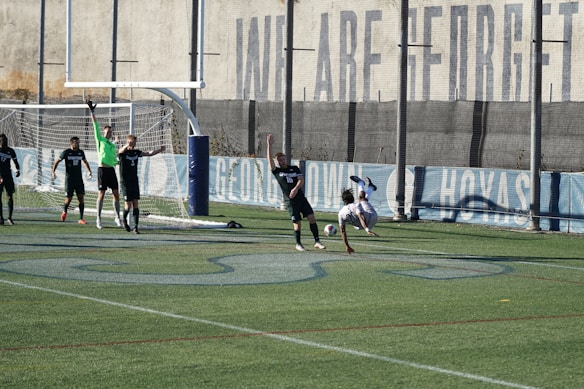 A soccer match on a field where players are in action near a goalpost. Several players in black uniforms are positioned in and around the goal area, while one player in white is executing an acrobatic move mid-air with a soccer ball nearby. The backdrop includes a tall wall with faded writing on it, and the field features green artificial turf with blue markings.