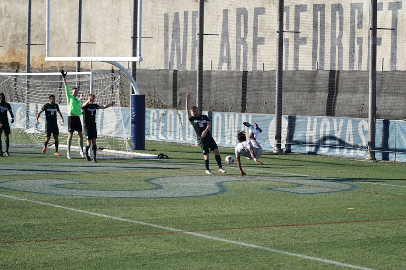 A soccer match on a field where players are in action near a goalpost. Several players in black uniforms are positioned in and around the goal area, while one player in white is executing an acrobatic move mid-air with a soccer ball nearby. The backdrop includes a tall wall with faded writing on it, and the field features green artificial turf with blue markings.