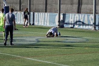A person is kneeling on a sports field, possibly in distress or exhaustion. There are several other individuals nearby, some standing and one holding a soccer ball. The field has markings typical of a sports ground, with a fence and a banner in the background.