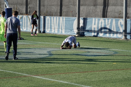 A person is kneeling on a sports field, possibly in distress or exhaustion. There are several other individuals nearby, some standing and one holding a soccer ball. The field has markings typical of a sports ground, with a fence and a banner in the background.