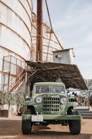 A vintage green jeep is parked in front of a large rusty industrial silo. The setting appears to be a rural or industrial area with metal structures and greenery climbing the building. The jeep has a license plate from Texas.
