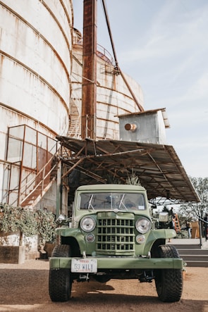 A vintage green jeep is parked in front of a large rusty industrial silo. The setting appears to be a rural or industrial area with metal structures and greenery climbing the building. The jeep has a license plate from Texas.