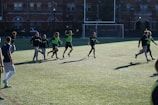 A group of men are playing a casual game of soccer on a grassy field. They are wearing sports attire, with some in black shirts and others wearing green vests. The lighting suggests late afternoon with long shadows cast on the field. In the background, there is a brick building and a soccer goal.