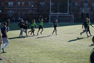 A group of men are playing a casual game of soccer on a grassy field. They are wearing sports attire, with some in black shirts and others wearing green vests. The lighting suggests late afternoon with long shadows cast on the field. In the background, there is a brick building and a soccer goal.