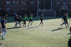 A group of men are playing a casual game of soccer on a grassy field. They are wearing sports attire, with some in black shirts and others wearing green vests. The lighting suggests late afternoon with long shadows cast on the field. In the background, there is a brick building and a soccer goal.