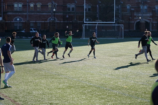 A group of men are playing a casual game of soccer on a grassy field. They are wearing sports attire, with some in black shirts and others wearing green vests. The lighting suggests late afternoon with long shadows cast on the field. In the background, there is a brick building and a soccer goal.