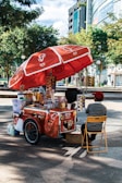 A street vendor's cart with a large red umbrella bearing logos and branding. The cart is filled with various items, including bottled drinks, snacks, and containers. The vendor, wearing a red cap and striped shirt, sits on a yellow folding chair beside the cart. The setting is outdoors, with trees and modern buildings in the background.