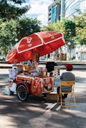 A street vendor's cart with a large red umbrella bearing logos and branding. The cart is filled with various items, including bottled drinks, snacks, and containers. The vendor, wearing a red cap and striped shirt, sits on a yellow folding chair beside the cart. The setting is outdoors, with trees and modern buildings in the background.