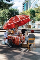 A street vendor's cart with a large red umbrella bearing logos and branding. The cart is filled with various items, including bottled drinks, snacks, and containers. The vendor, wearing a red cap and striped shirt, sits on a yellow folding chair beside the cart. The setting is outdoors, with trees and modern buildings in the background.