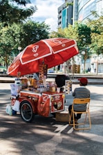 A street vendor's cart with a large red umbrella bearing logos and branding. The cart is filled with various items, including bottled drinks, snacks, and containers. The vendor, wearing a red cap and striped shirt, sits on a yellow folding chair beside the cart. The setting is outdoors, with trees and modern buildings in the background.