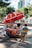 A street vendor's cart with a large red umbrella bearing logos and branding. The cart is filled with various items, including bottled drinks, snacks, and containers. The vendor, wearing a red cap and striped shirt, sits on a yellow folding chair beside the cart. The setting is outdoors, with trees and modern buildings in the background.