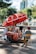A street vendor's cart with a large red umbrella bearing logos and branding. The cart is filled with various items, including bottled drinks, snacks, and containers. The vendor, wearing a red cap and striped shirt, sits on a yellow folding chair beside the cart. The setting is outdoors, with trees and modern buildings in the background.
