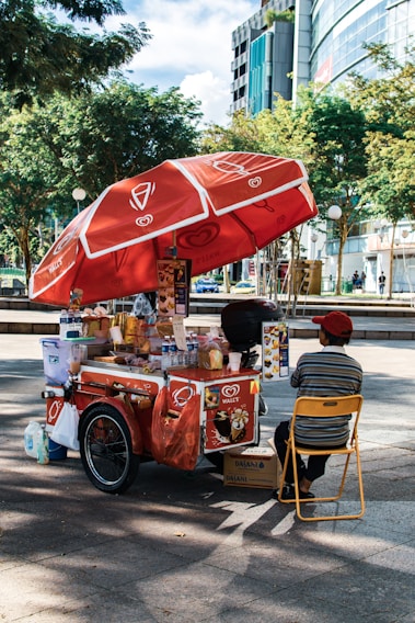 A street vendor's cart with a large red umbrella bearing logos and branding. The cart is filled with various items, including bottled drinks, snacks, and containers. The vendor, wearing a red cap and striped shirt, sits on a yellow folding chair beside the cart. The setting is outdoors, with trees and modern buildings in the background.